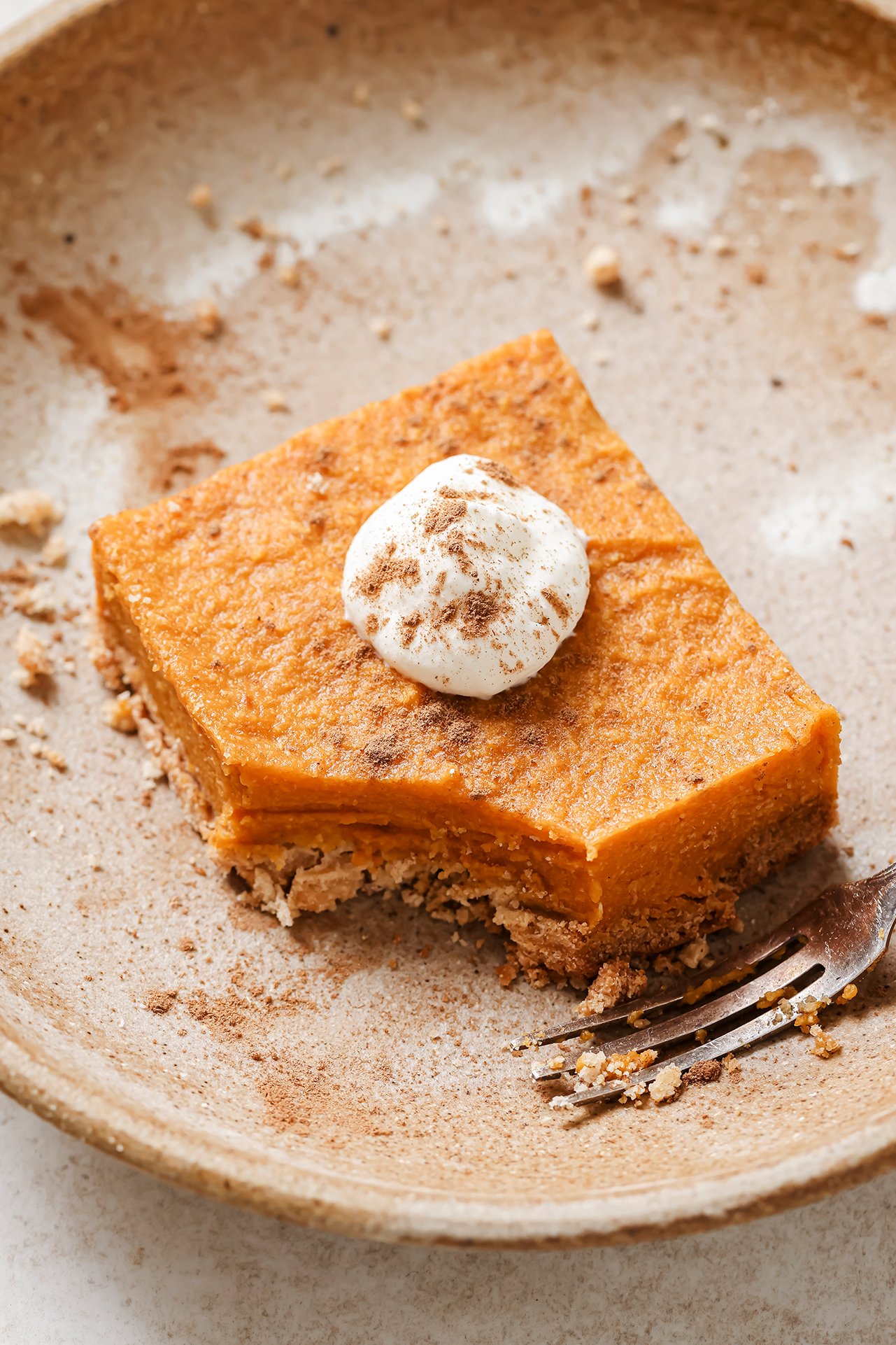 One pumpkin pie bar on a plate topped with coconut cream and cinnamon.