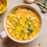 A bowl of broccoli cheese noodle soup in a bowl with a handle and a spoon to the side.