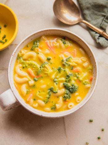 A bowl of broccoli cheese noodle soup in a bowl with a handle and a spoon to the side.
