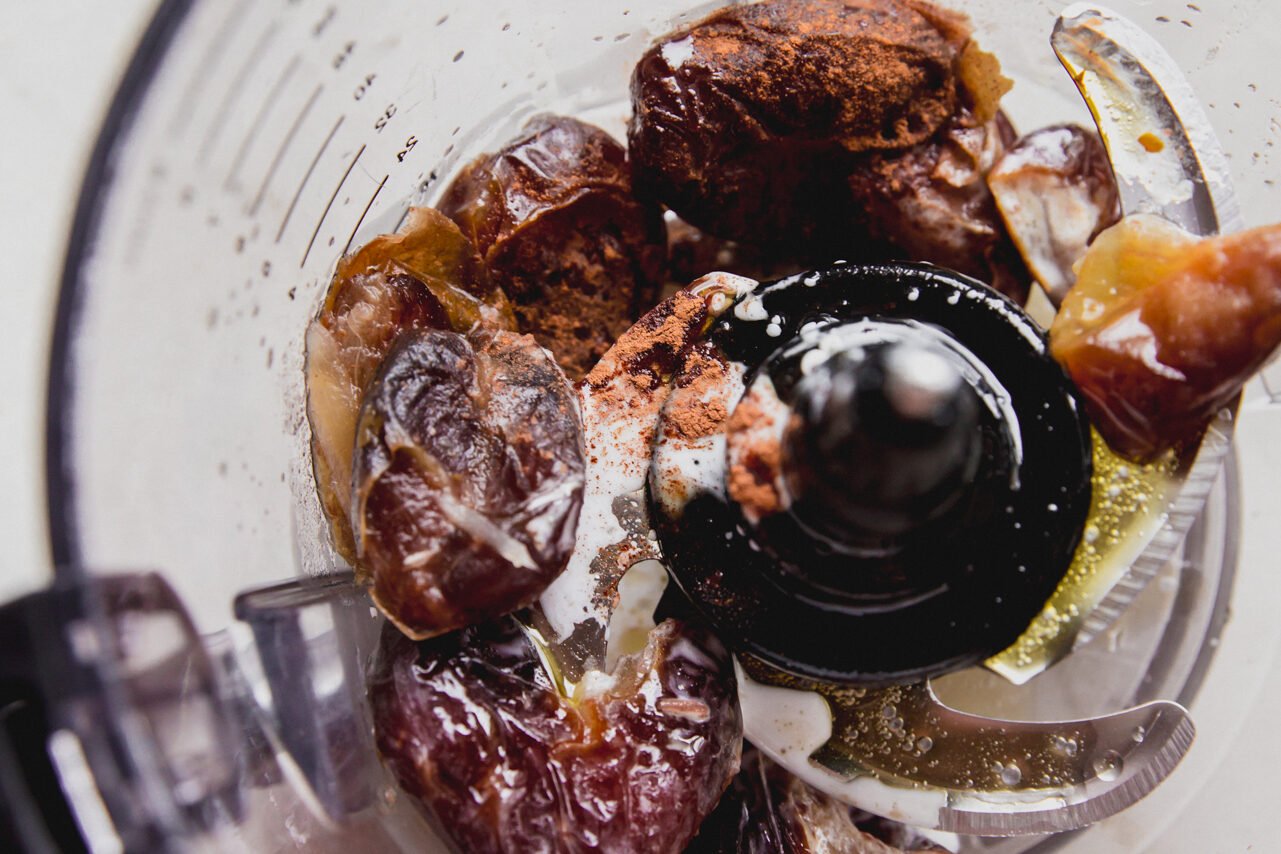 A food processor blending dates, coconut oil, ghee, and cinnamon.