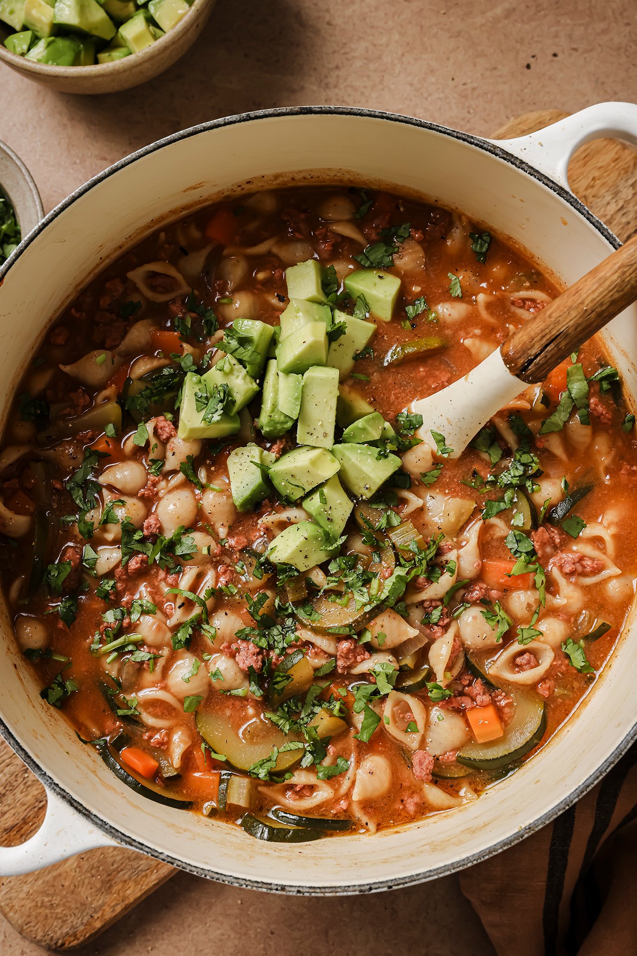 Conchitas con carne soup in a large white dutch oven, topped with avocado and cilantro, and avocado on the side.