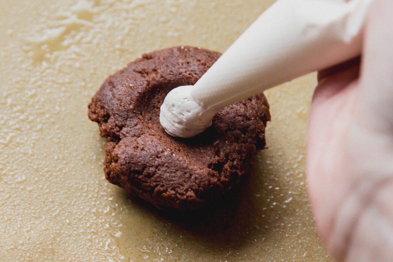 A hand pipping marshmallow fluff into a cookie on a piece of parchment paper.