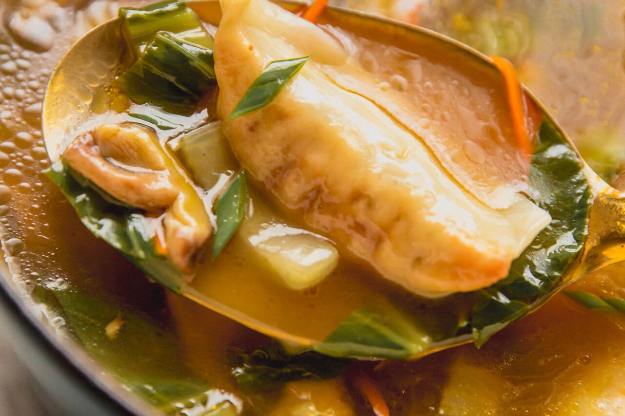 A metal serving spoon lifting up a posticker with broth and vegetables.