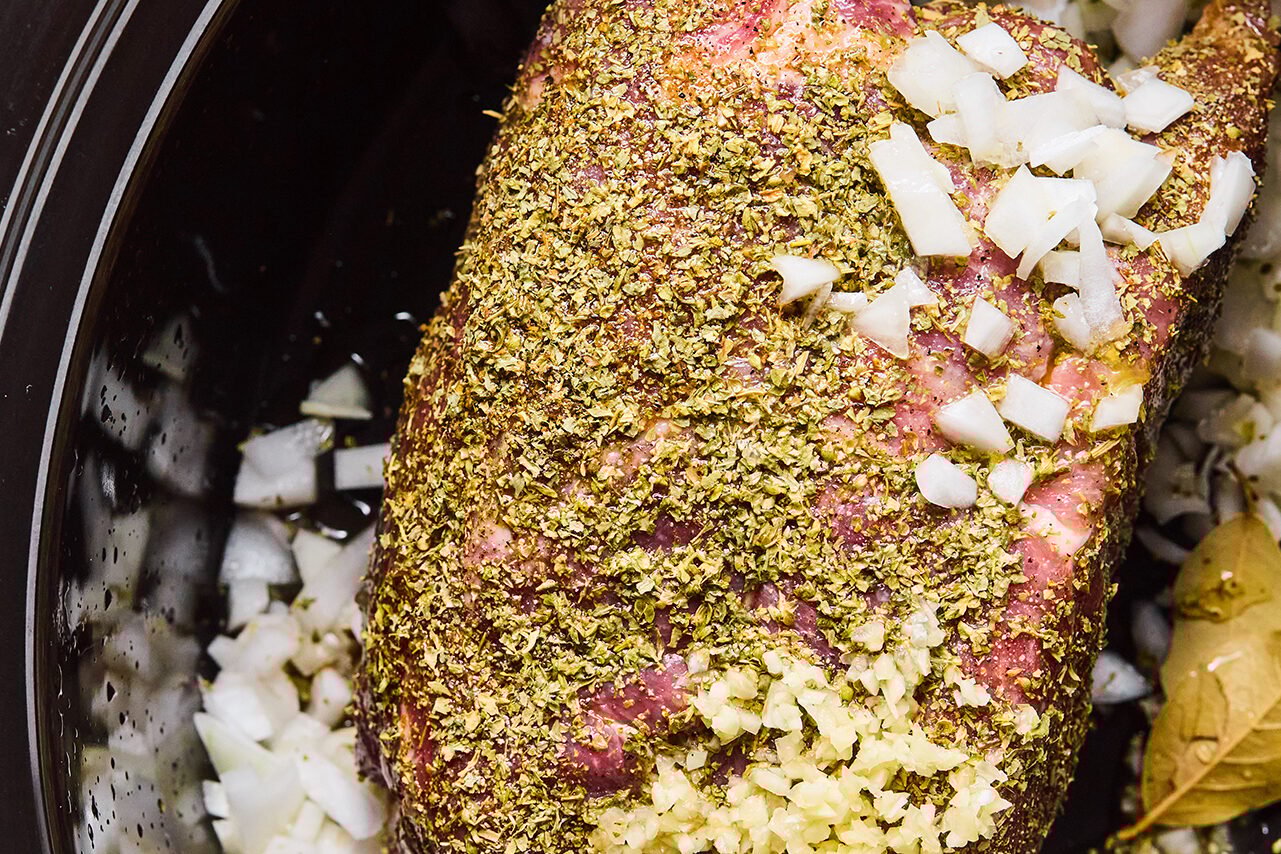 Pork shoulder in a slow cooker coated in a spice rub, with diced onion, minced garlic, and juice.