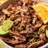 Slow cooker carnitas shredded in a bowl and topped with sliced lime, orange and chopped cilantro.