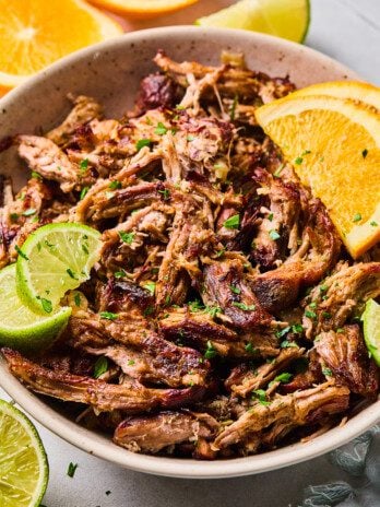 Slow cooker carnitas shredded in a bowl and topped with sliced lime, orange and chopped cilantro.