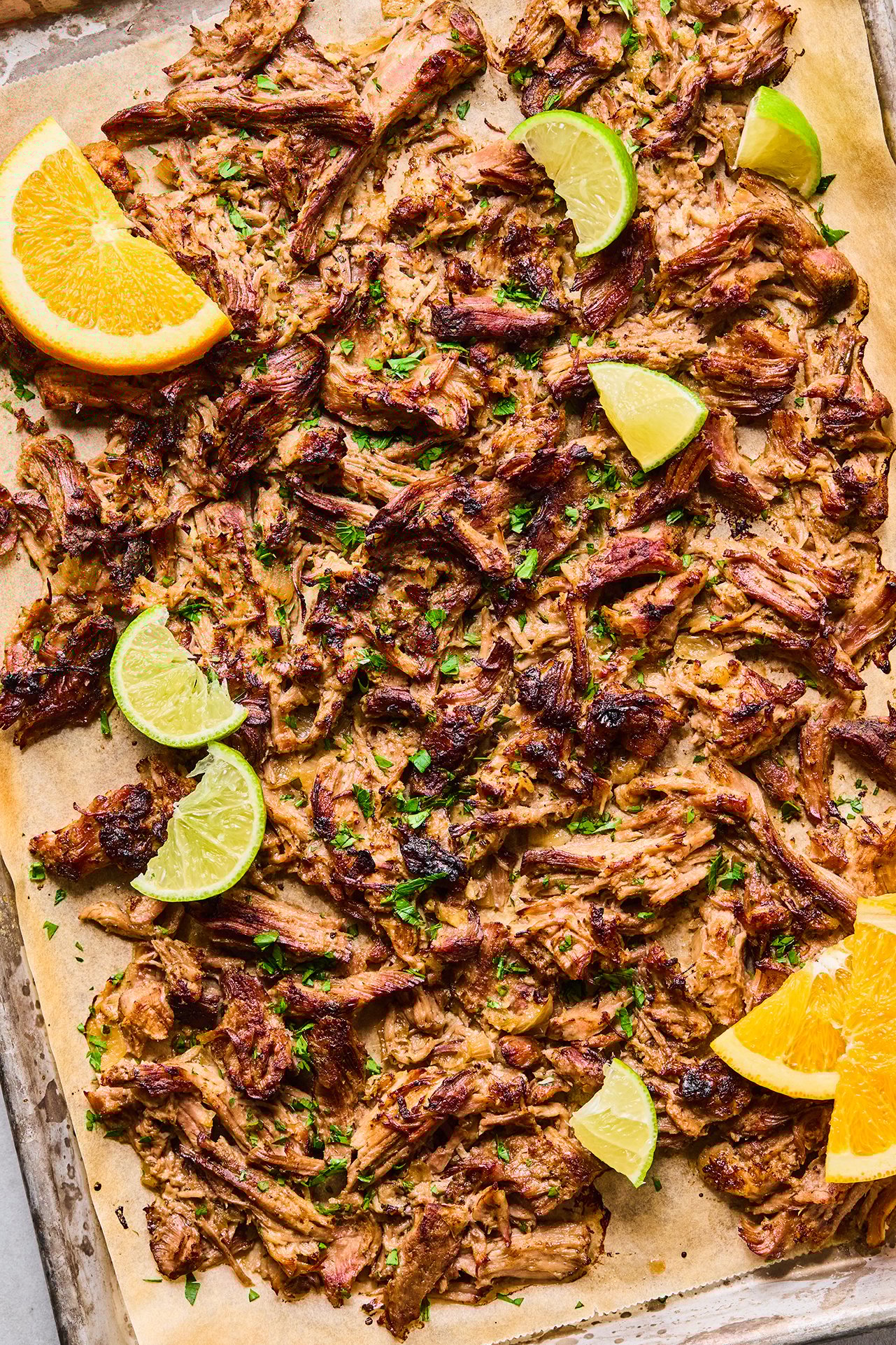 Crispy carnitas on a parchment lined baking sheet topped with chopped cilantro with lime wedges and orange wedges.