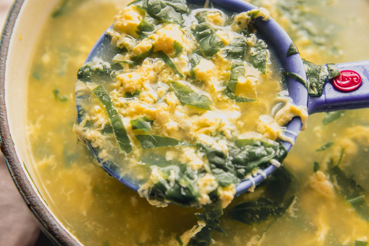 A measuring cup holding a portion of stracciatella soup above the pot of it.