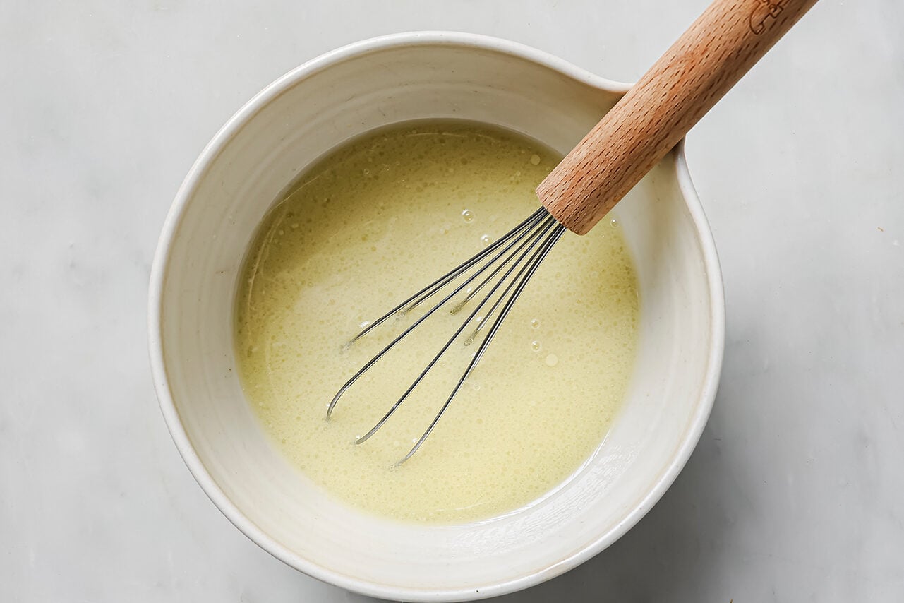 A whisk combining coconut milk, arrowroot starch, lemon juice and nutritional yeast in a white bowl.