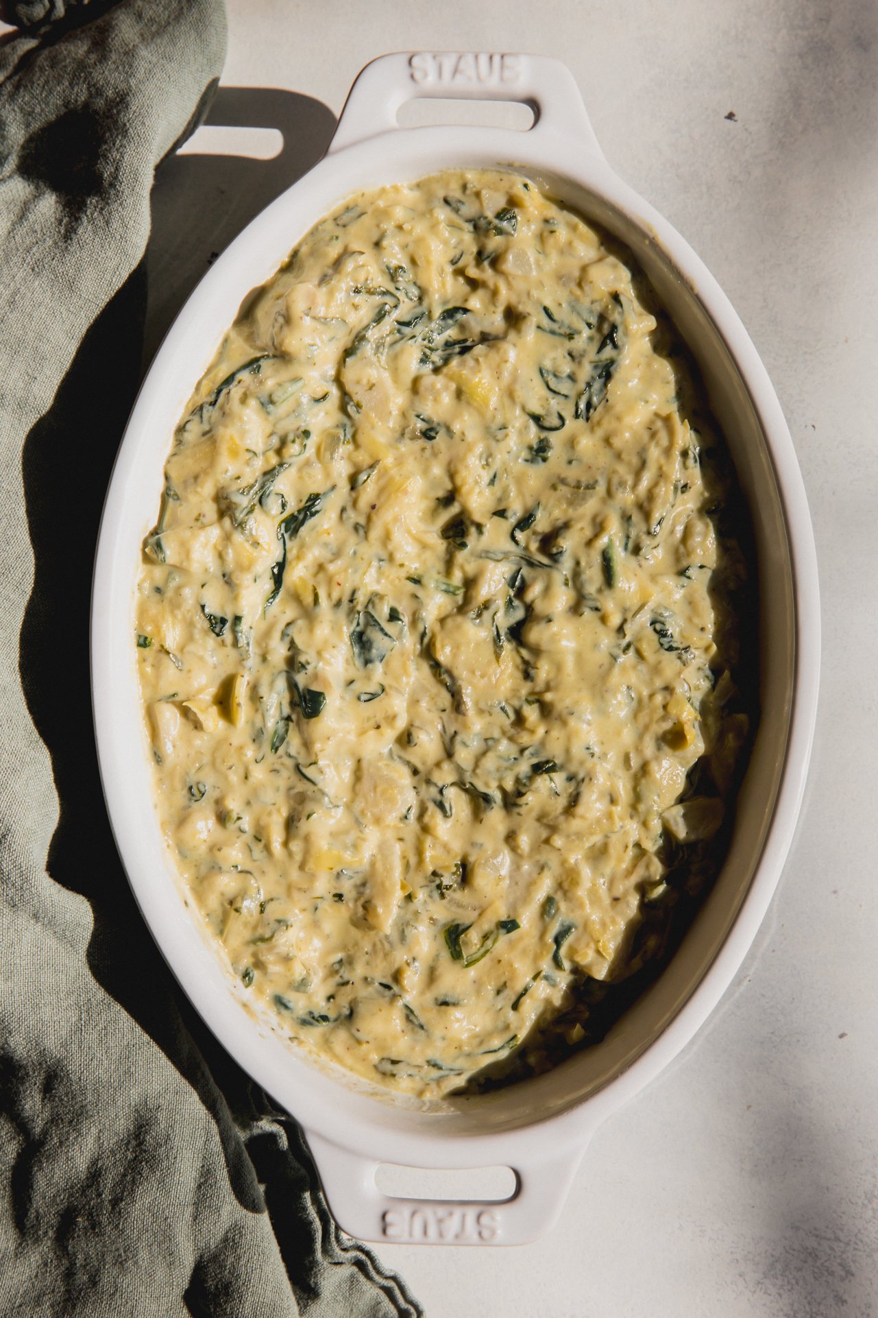 A white baking dish filled with spinach and artichoke dip after baking with a green dish towel next to it.