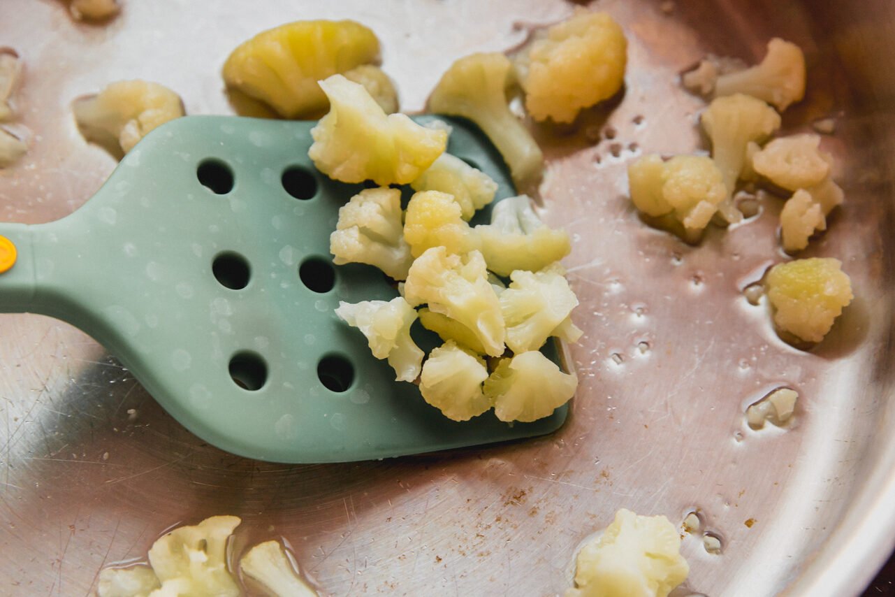 Cauliflower florets being scooped by a spatula from a pan after steaming.
