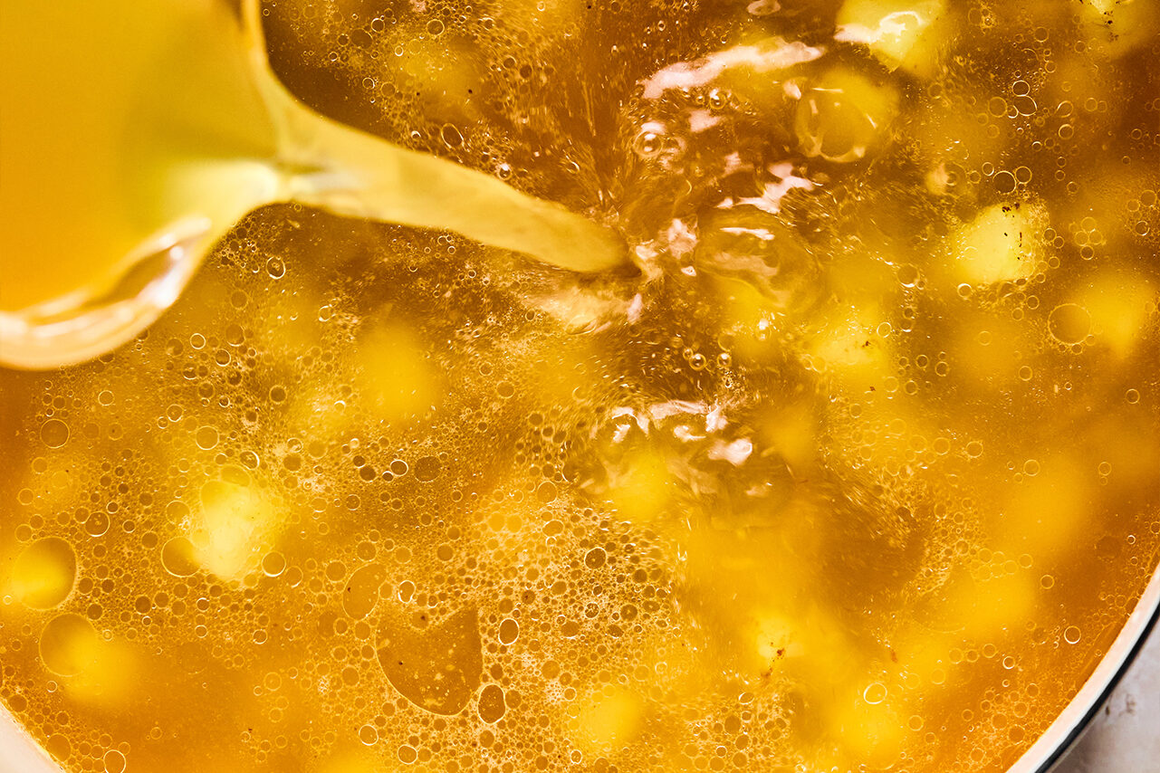 Broth and sweet potatoes in a pan with broth mixed with arrowroot starch being poured in.