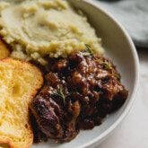 French onion short ribs topped with fresh thyme in a bowl with mashed sweet potato and cheese toast.