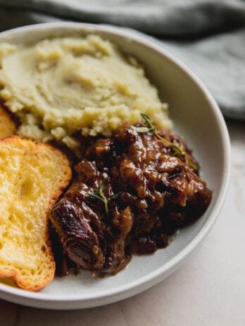 French onion short ribs topped with fresh thyme in a bowl with mashed sweet potato and cheese toast.