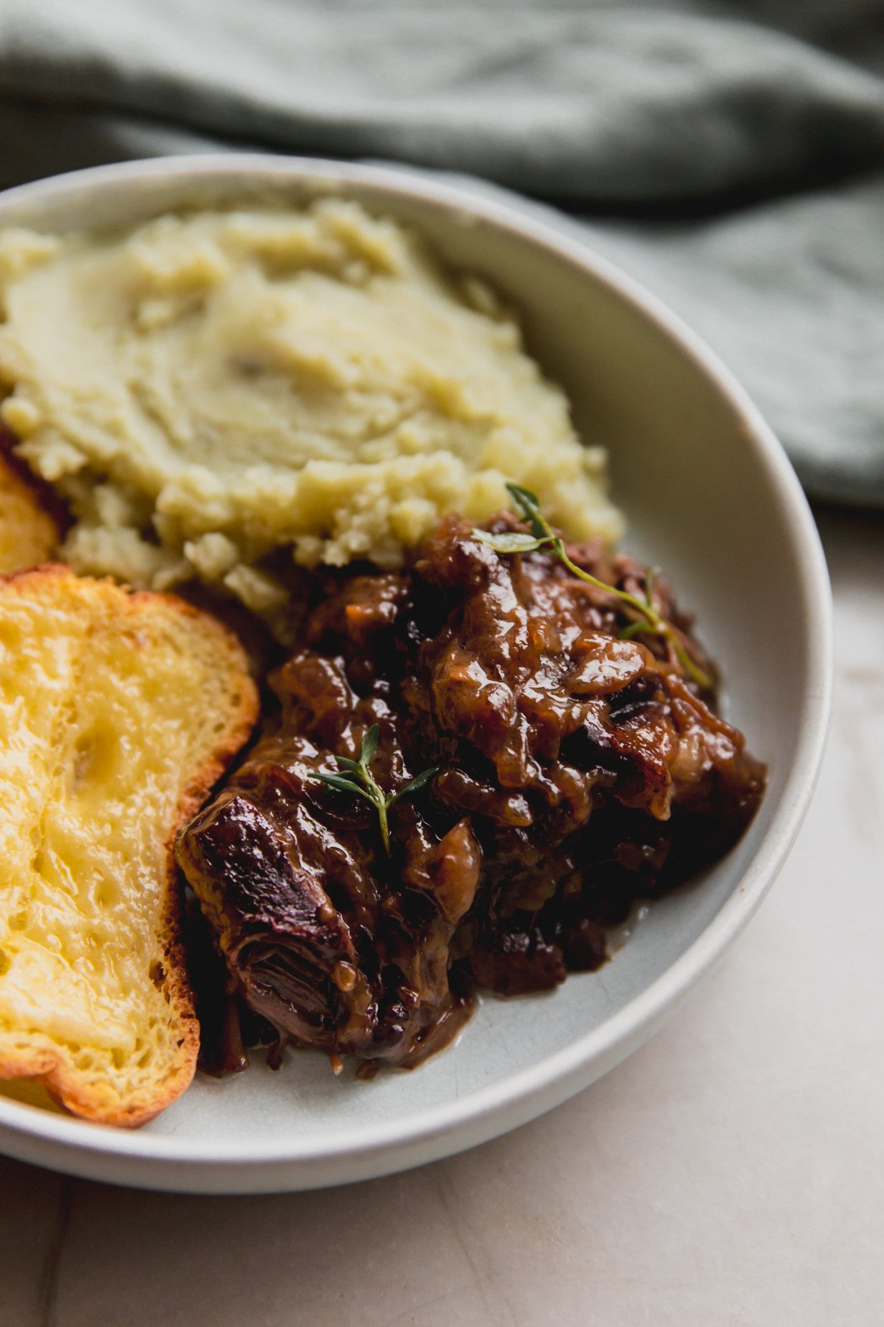 French onion short ribs topped with fresh thyme in a bowl with mashed sweet potato and cheese toast.