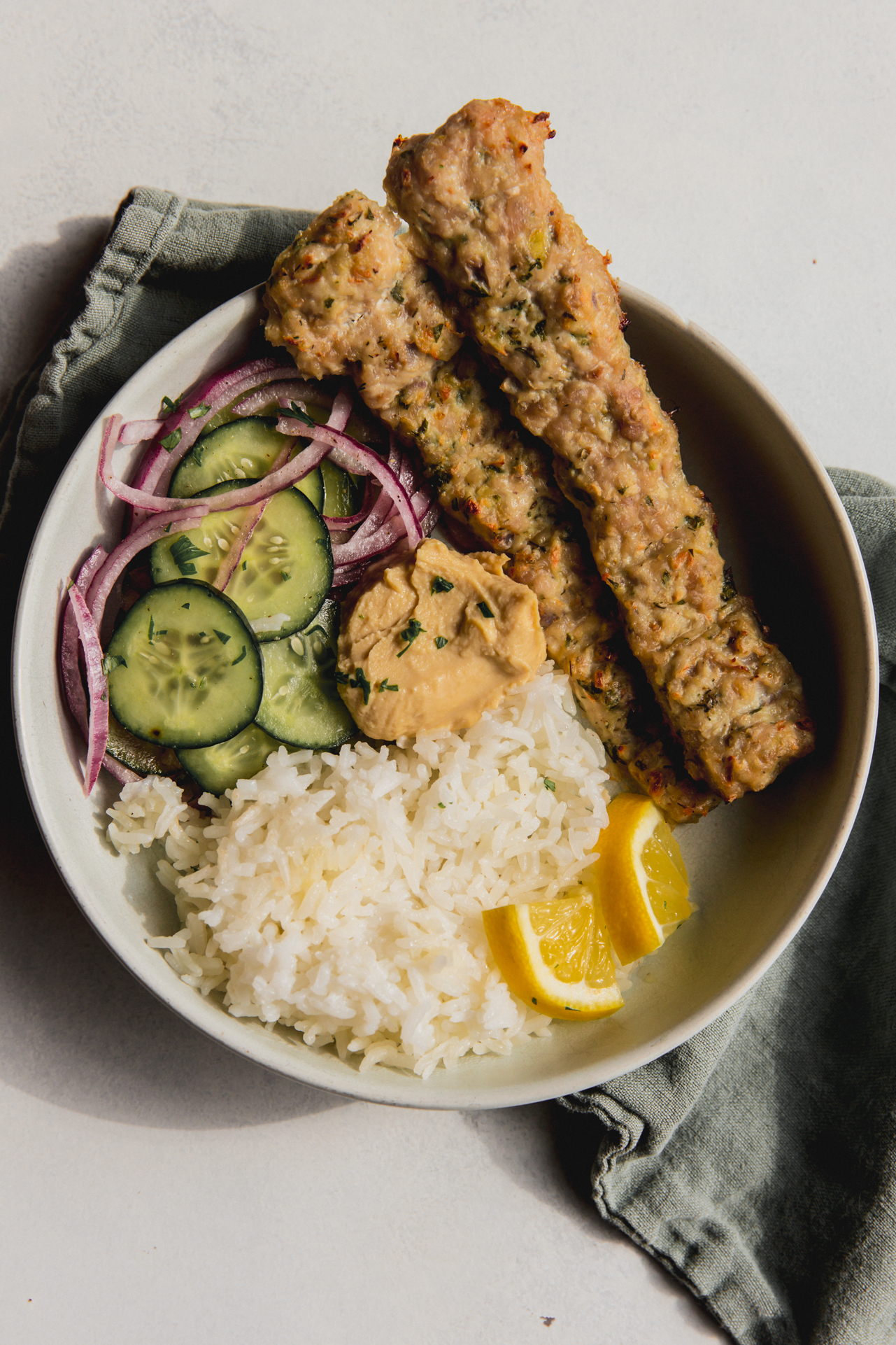 Two sheet pan kebabs in a bowl with cucumber salad, white rice and sliced lemon wedges.