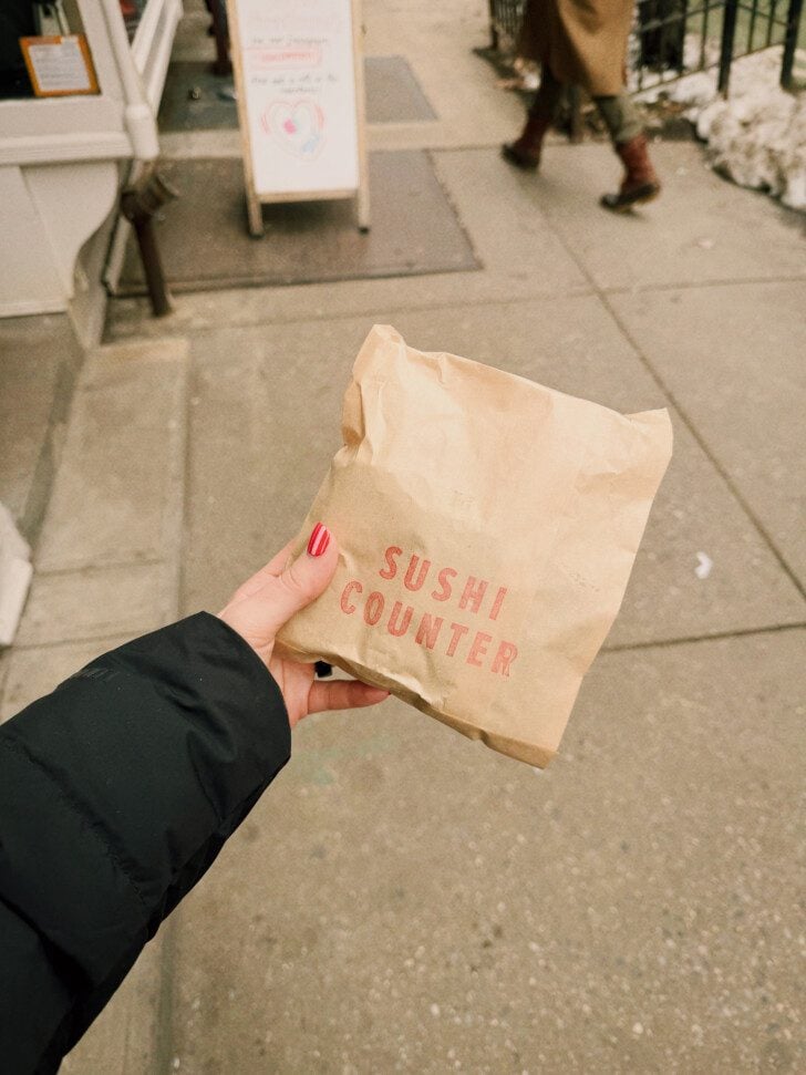 A hand holding a bag that says "sushi counter" with the side walk in the background