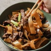 Gluten free beef chow fun in a shallow green bowl with noodles being pulled up with chopsticks.