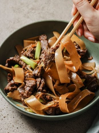 Gluten free beef chow fun in a shallow green bowl with noodles being pulled up with chopsticks.