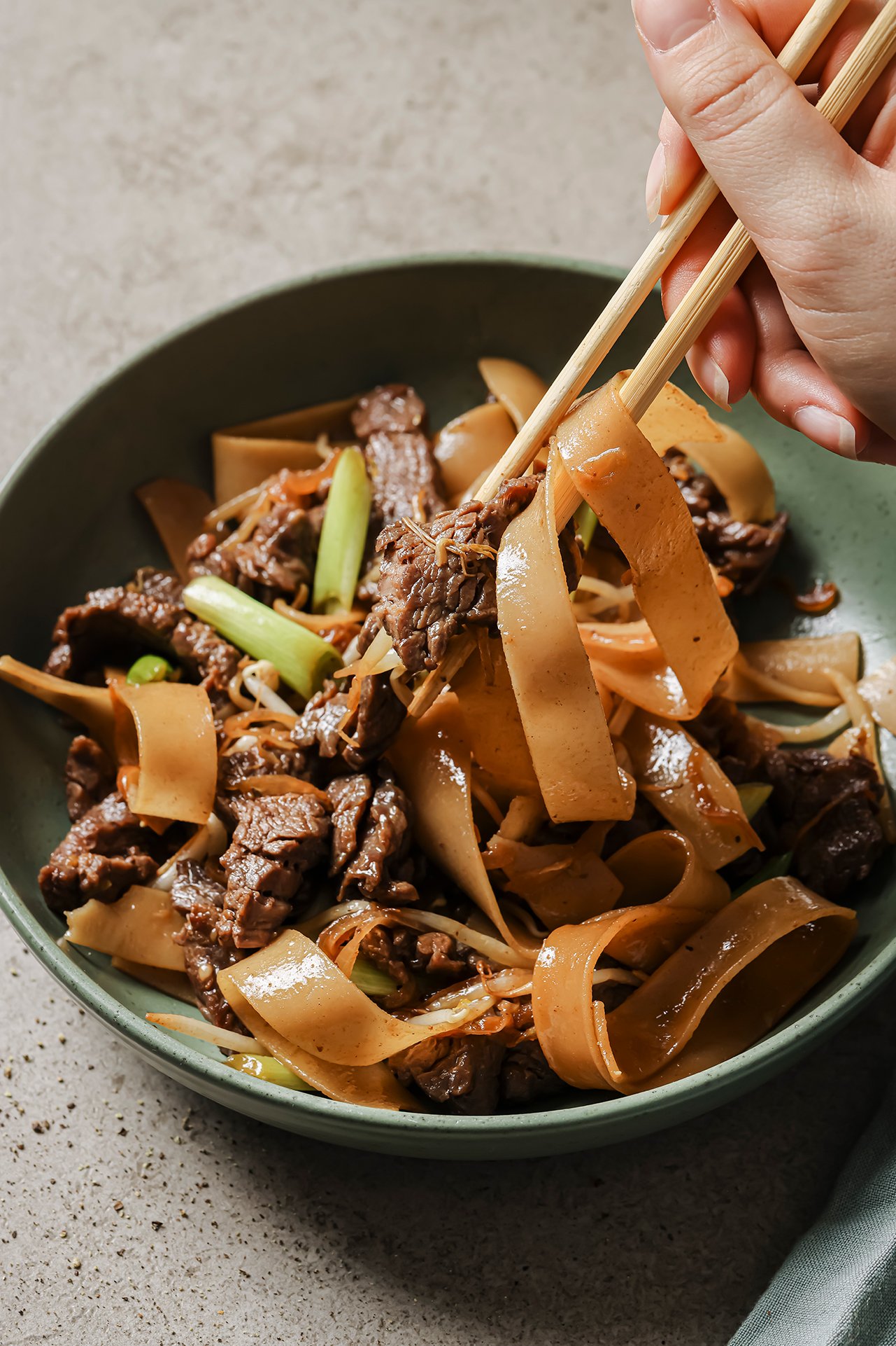 Gluten free beef chow fun in a shallow green bowl with noodles being pulled up with chopsticks.