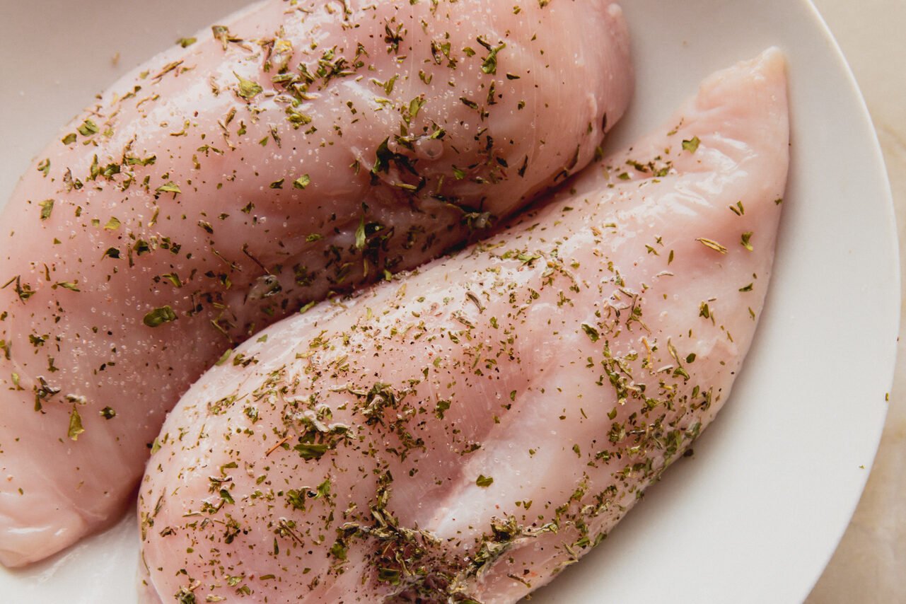 Raw chicken breasts on a white plate with seasoning on it.