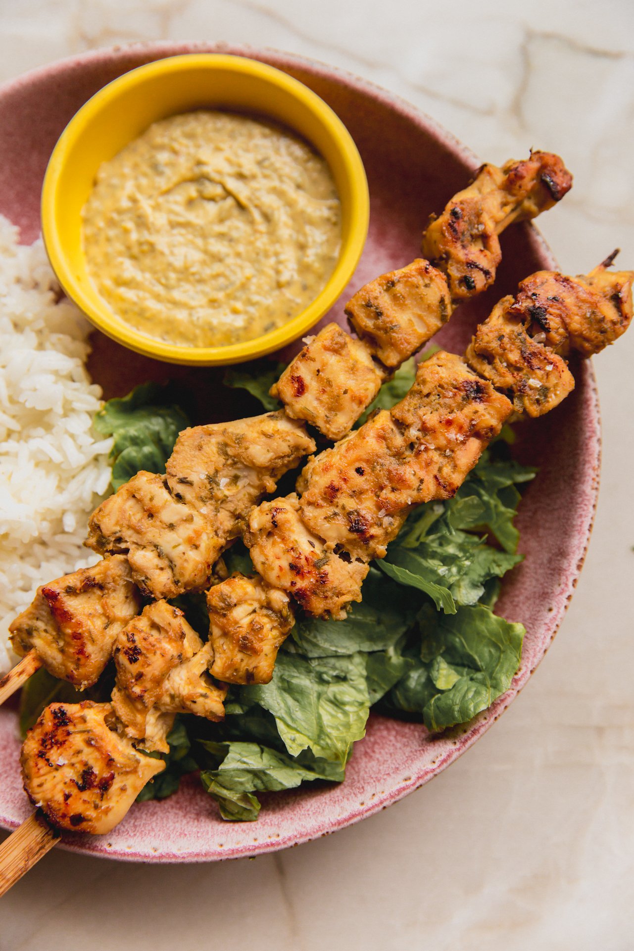 Garlic and herb chicken skewers in a bowl with dip on the side, rice and lettuce.