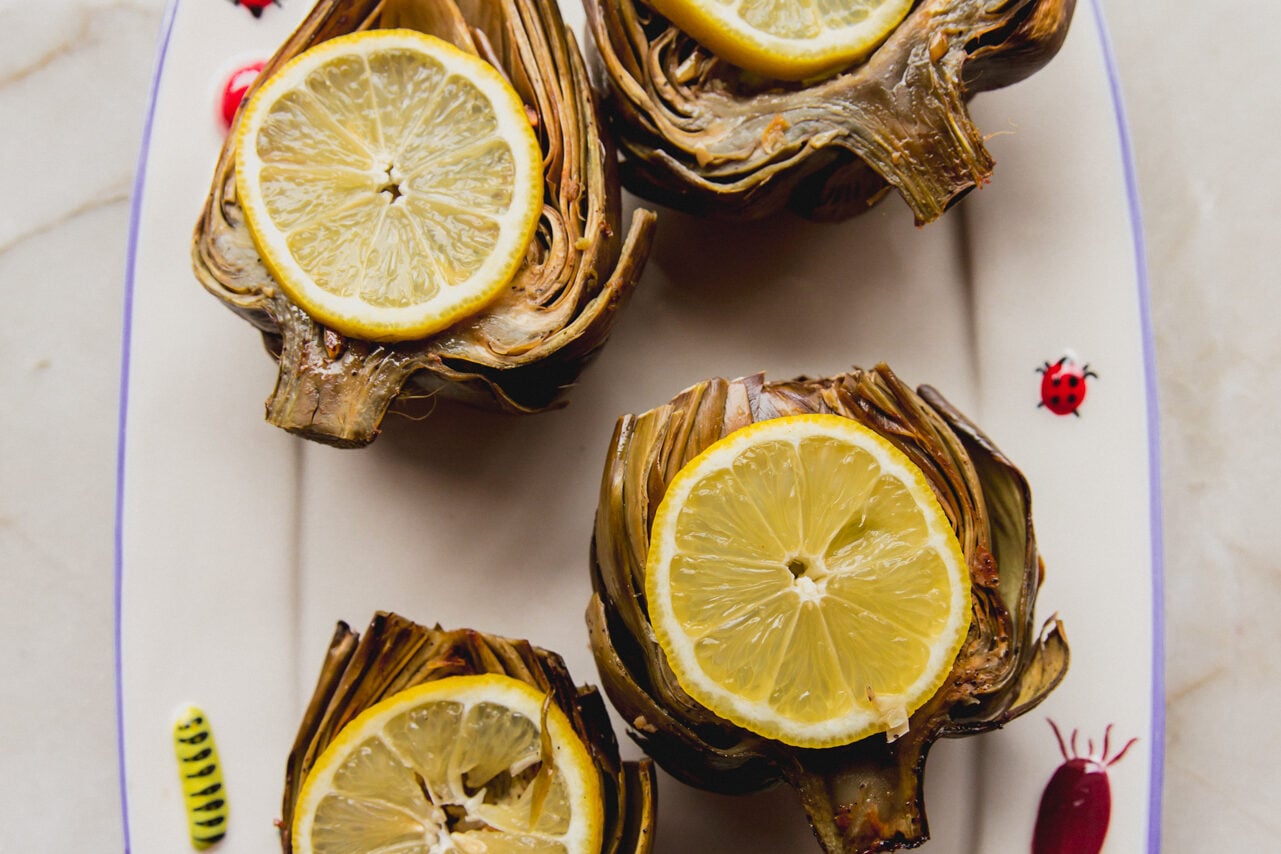 Four oven baked artichokes topped with sliced lemon and sitting on a platter.