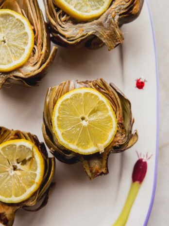 Four oven baked artichokes topped with sliced lemon and sitting on a platter.