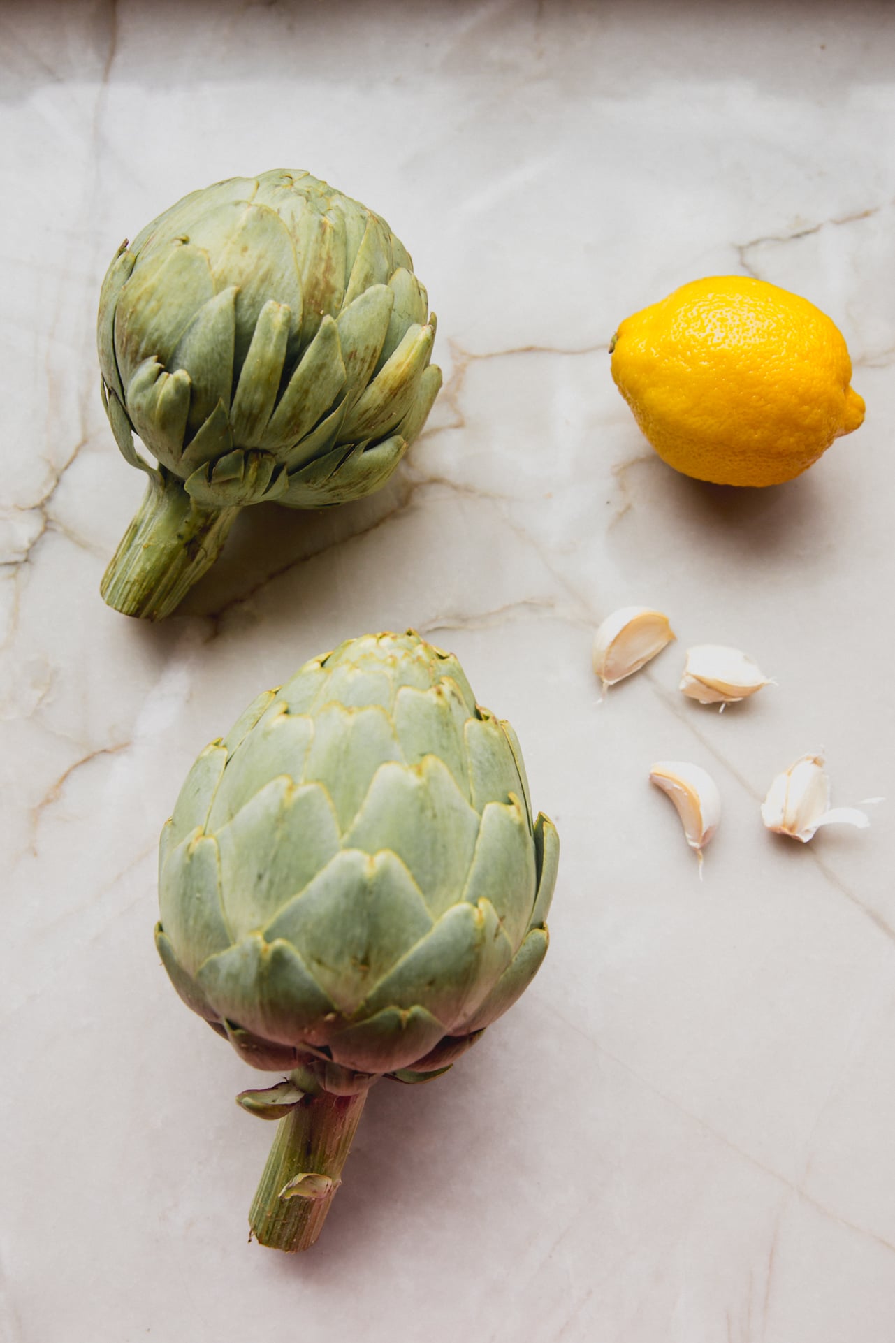 Two artichokes, a lemon and garlic cloves on a marble counter top.