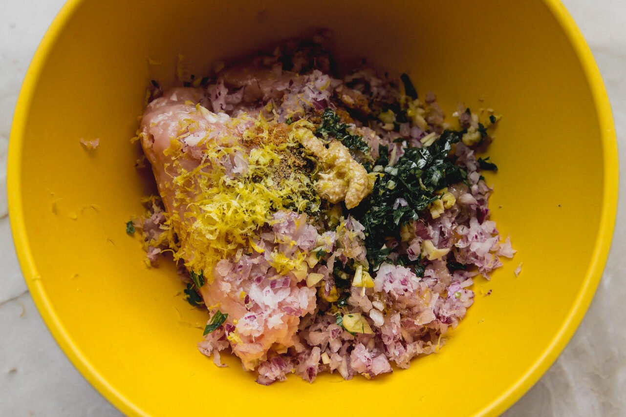 A yellow mixing bowl with ground chicken, seasonings, lemon zest, and red onion before being mixed together.