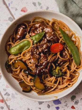 Steak noodle stir fry in a bowl with veggies and on a colorful surface.