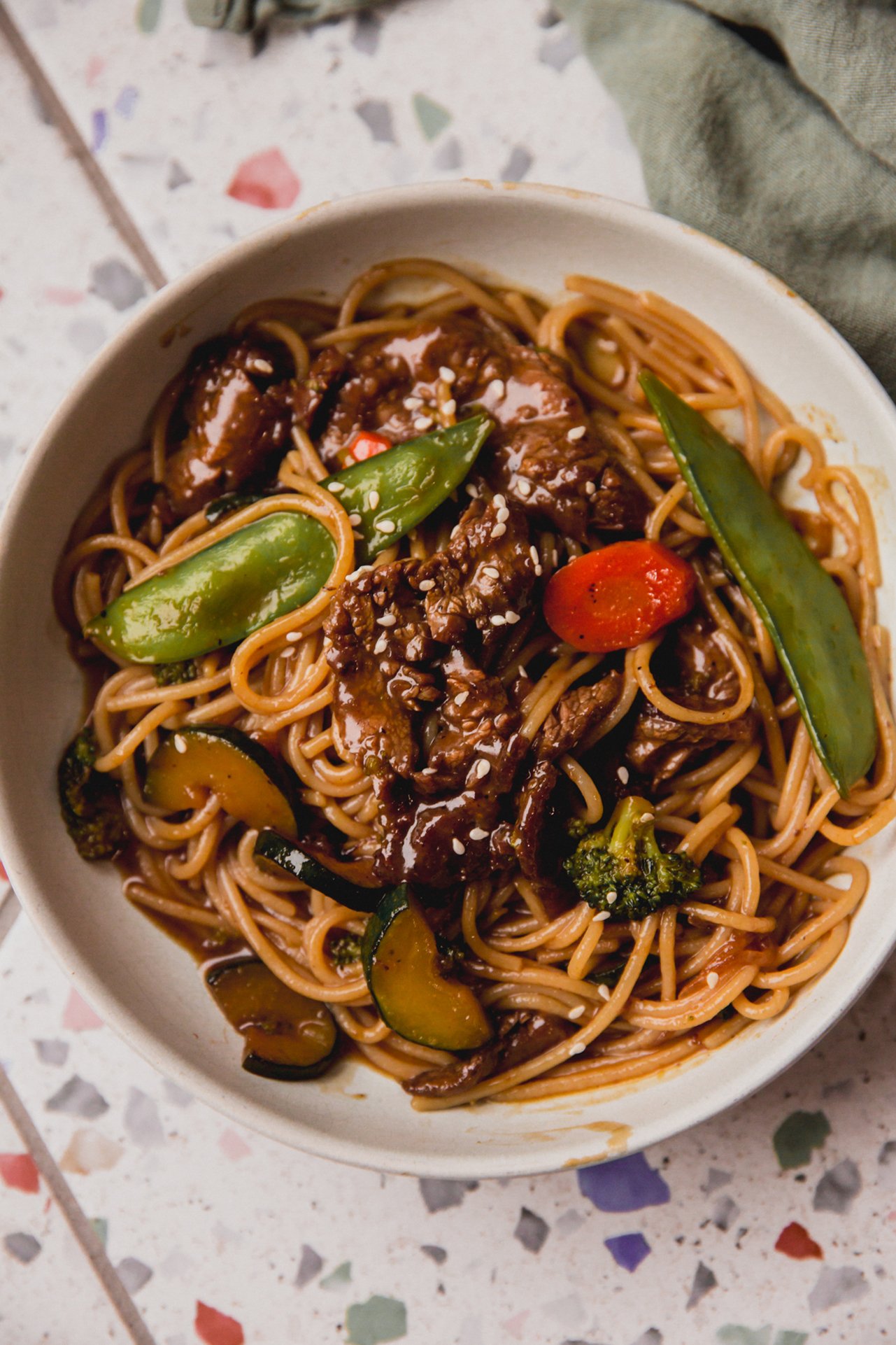 Steak noodle stir fry in a bowl with veggies and on a colorful surface.
