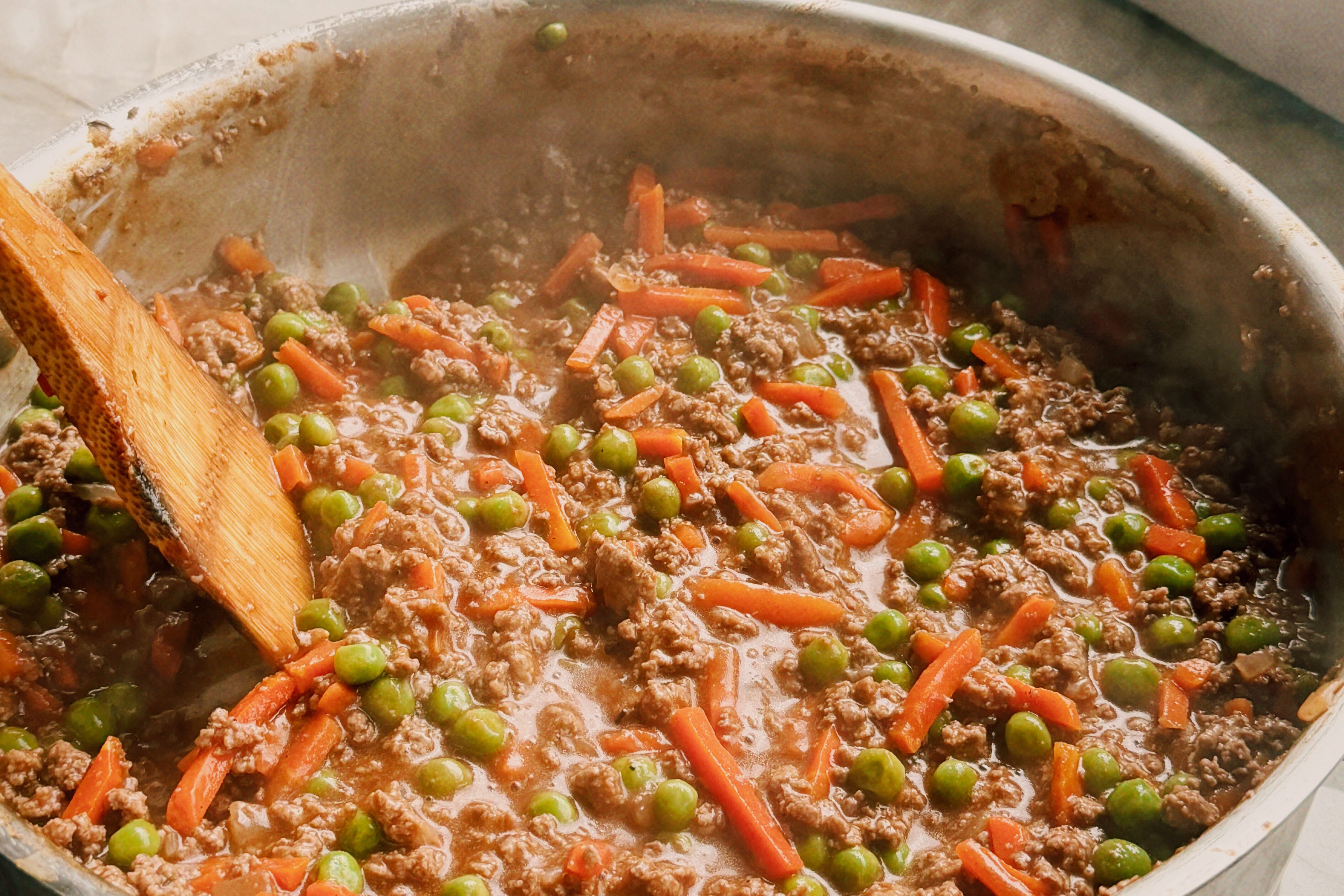A saute pan with ground beef, peas and carrots, and a wooden spoon stirring it.
