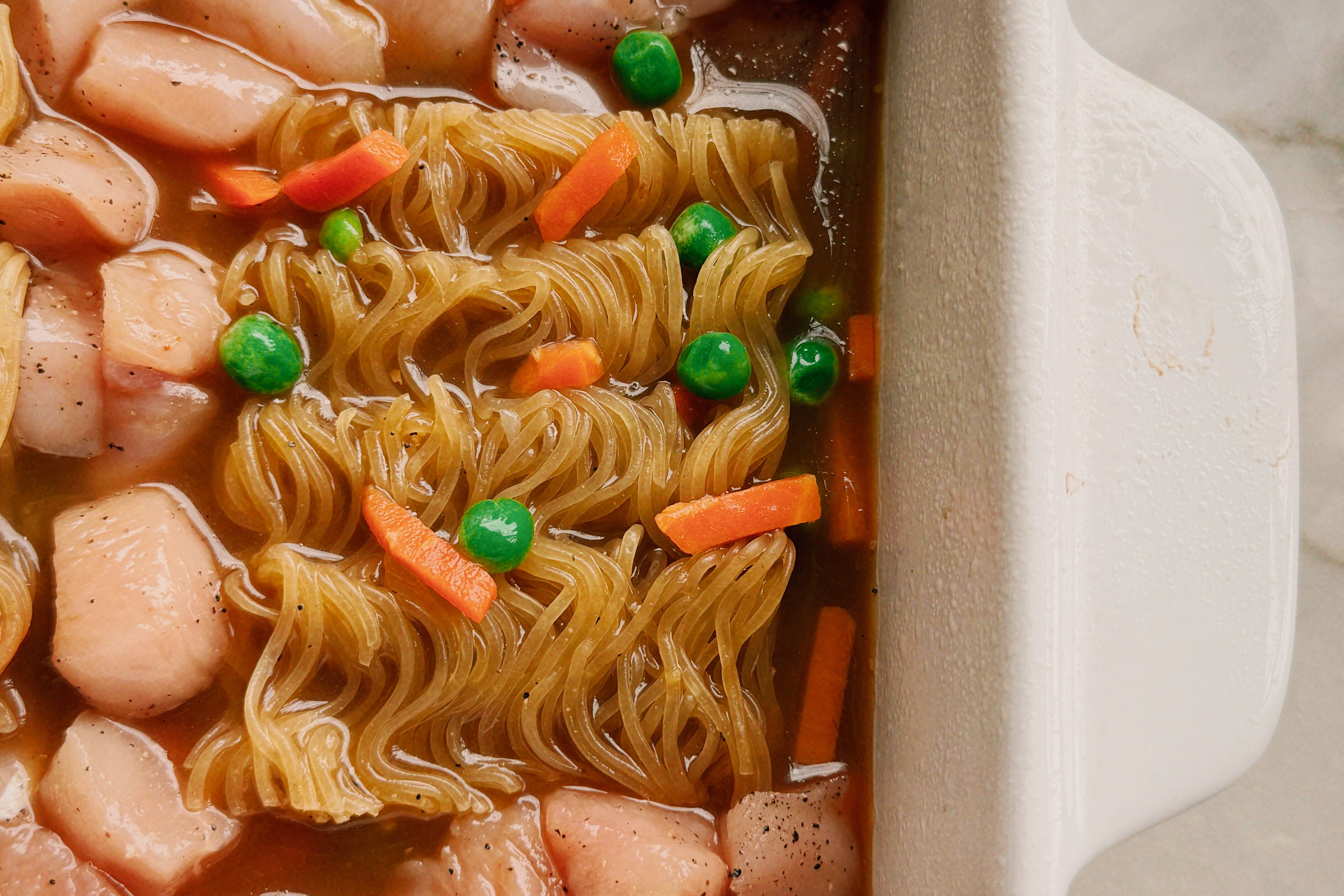 Uncooked ramen brick, uncooked chicken, carrots and peas, and sauce in a casserole dish.