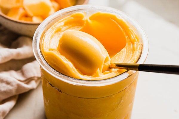 Mango ninja creami sorbet being scooped with a spoon, with frozen mango in a bowl in the background.