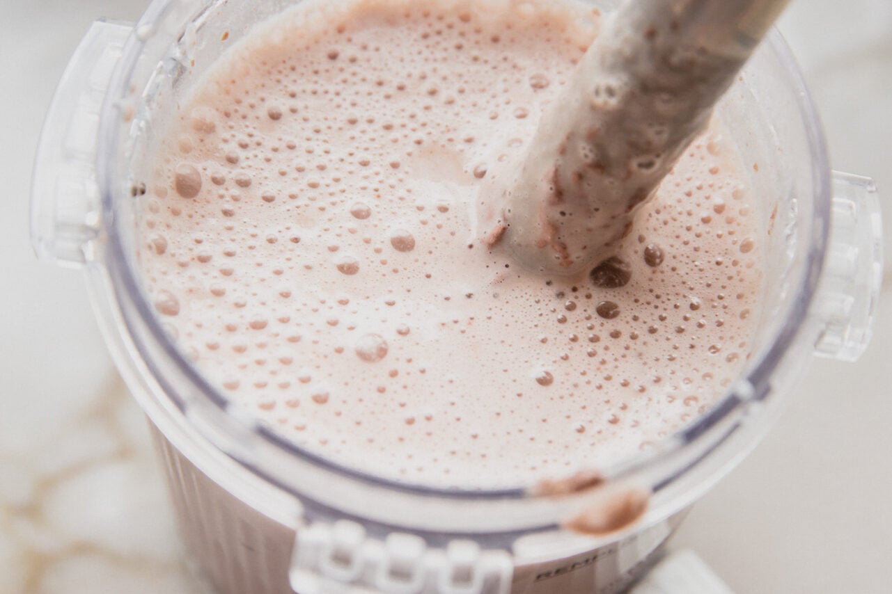 The pint container of a creami filled with all the ingredients and an immersion blender mixing it.