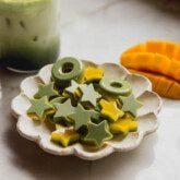 Star and ring shaped two tone mango and matcha gummies on a plate with a sliced mango and matcha latte in the background.
