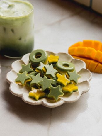 Star and ring shaped two tone mango and matcha gummies on a plate with a sliced mango and matcha latte in the background.