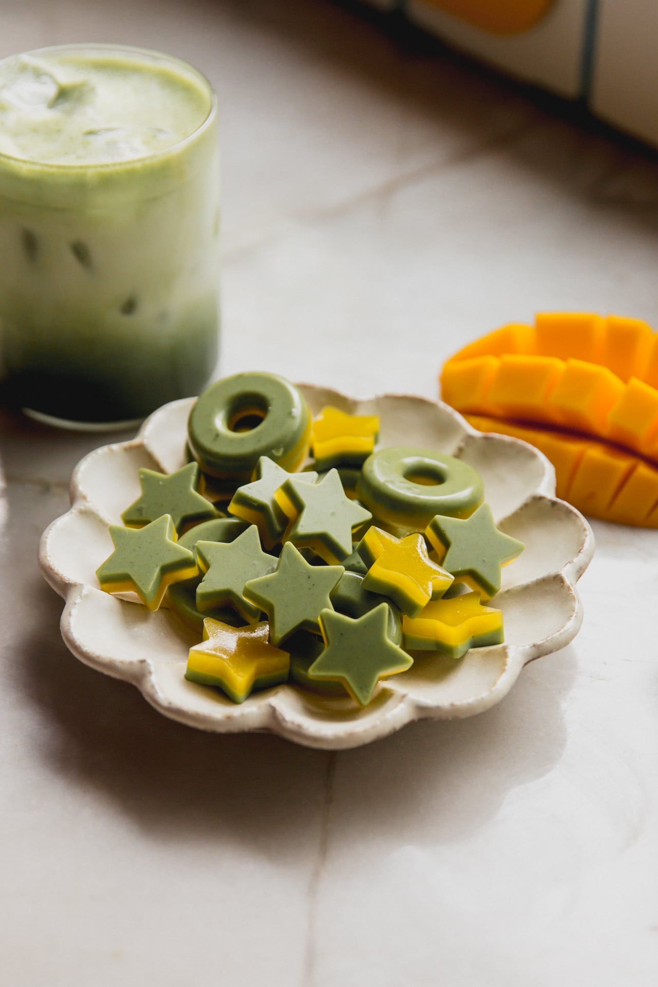 Star and ring shaped two tone mango and matcha gummies on a plate with a sliced mango and matcha latte in the background.