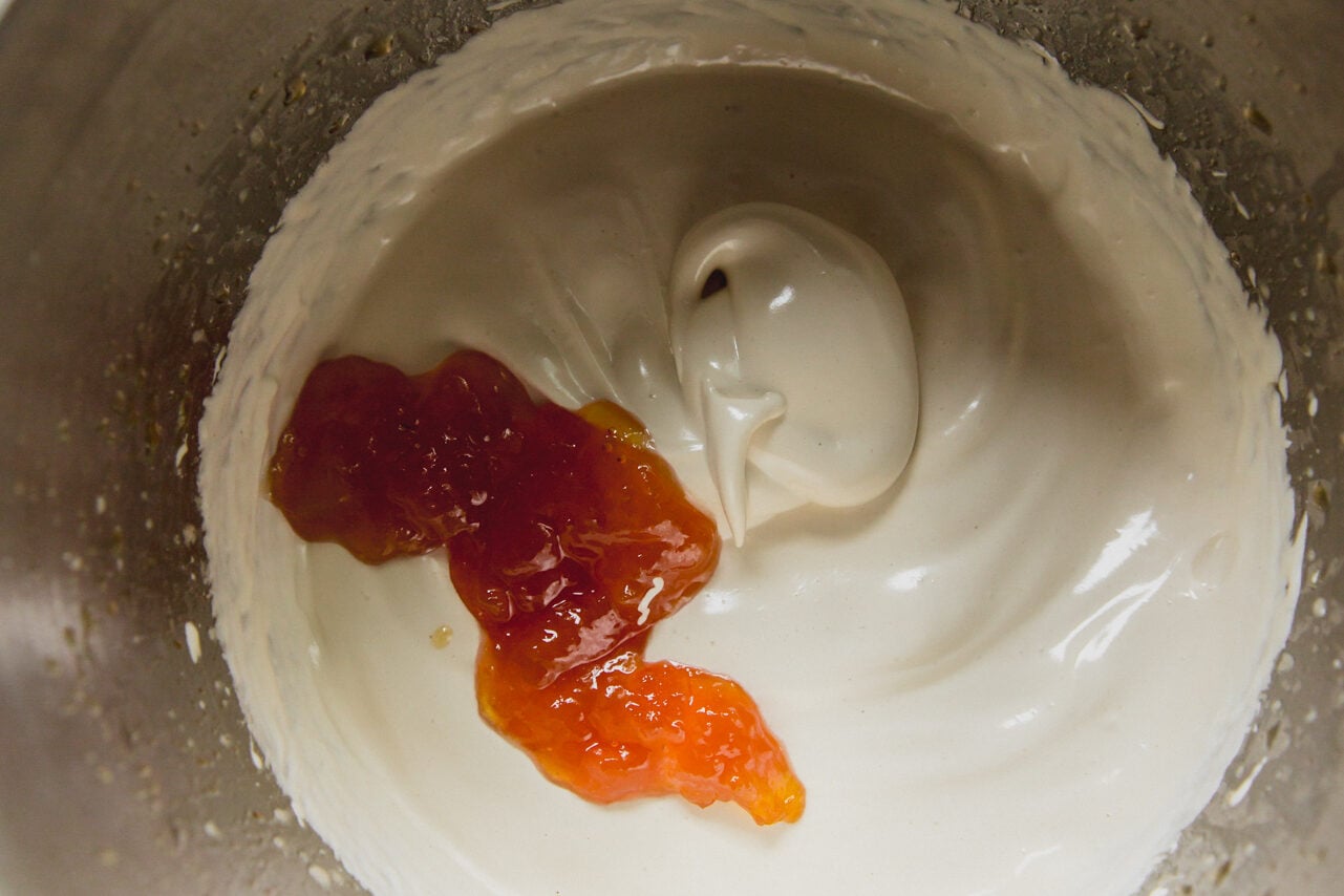 The stainless steel bowl of a stand mixer with marshmallow whip and peach jam being added to it.