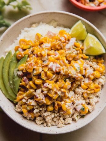 A bowl of rice and ground chicken topped with street corn, avocado and lime.