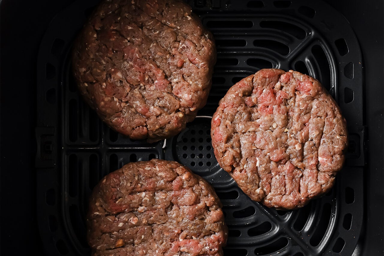 Three burgers uncooked in an air fryer basket.