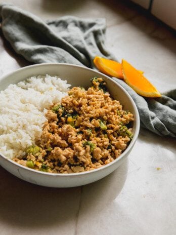 Orange ground chicken bowls in a bowl with rice and orange slices in the backround.
