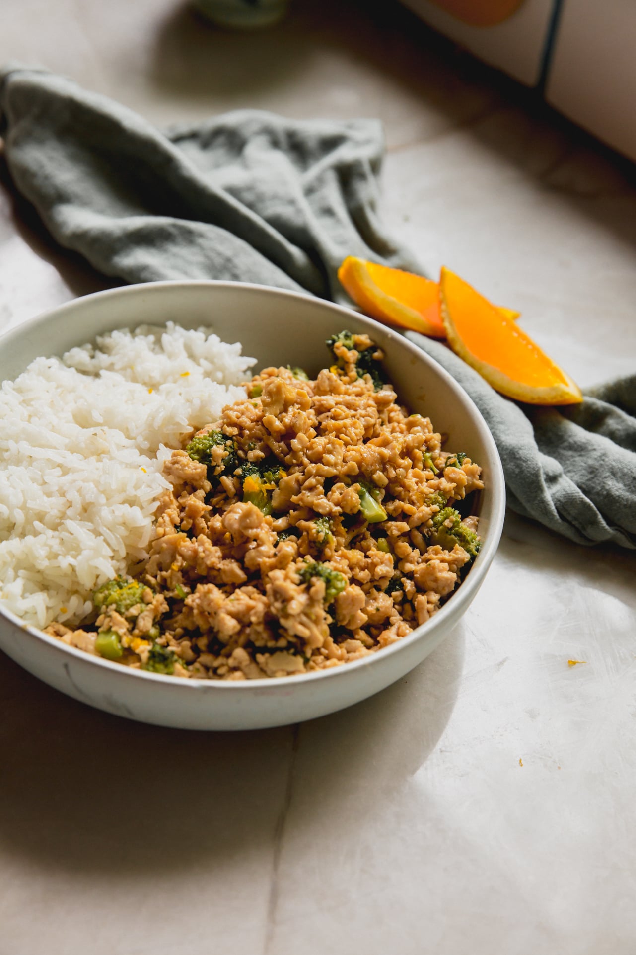 Orange ground chicken bowls in a bowl with rice and orange slices in the backround.