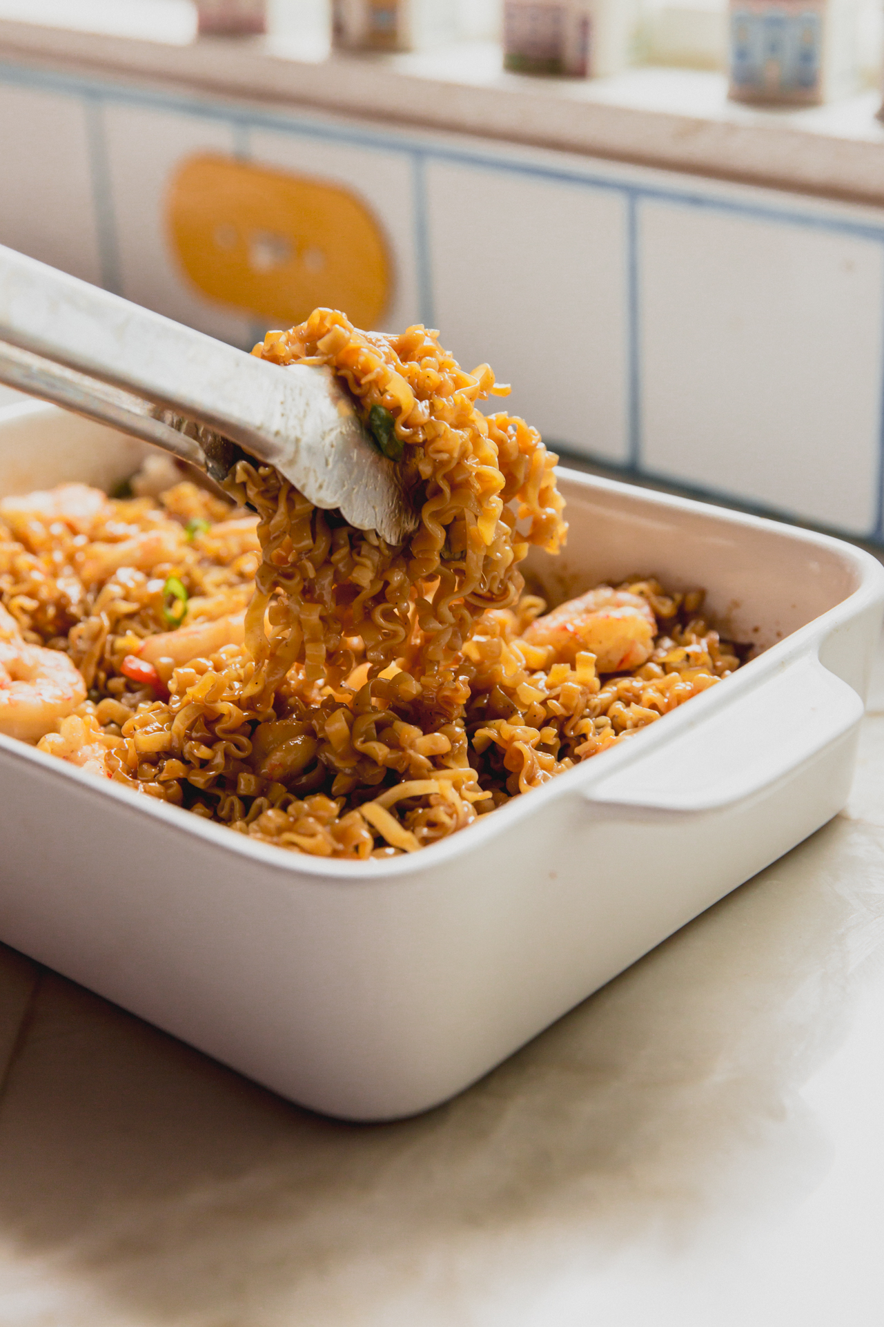 A pair of metal thongs lifting some of the shrimp ramen noodle bake out of the white ceramic dish it was baked it.