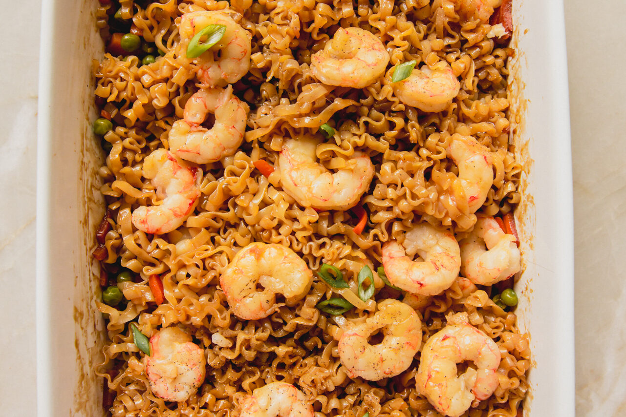 A white casserole dish with cooked shrimp on top of a ramen and vegetable sauce mixture.
