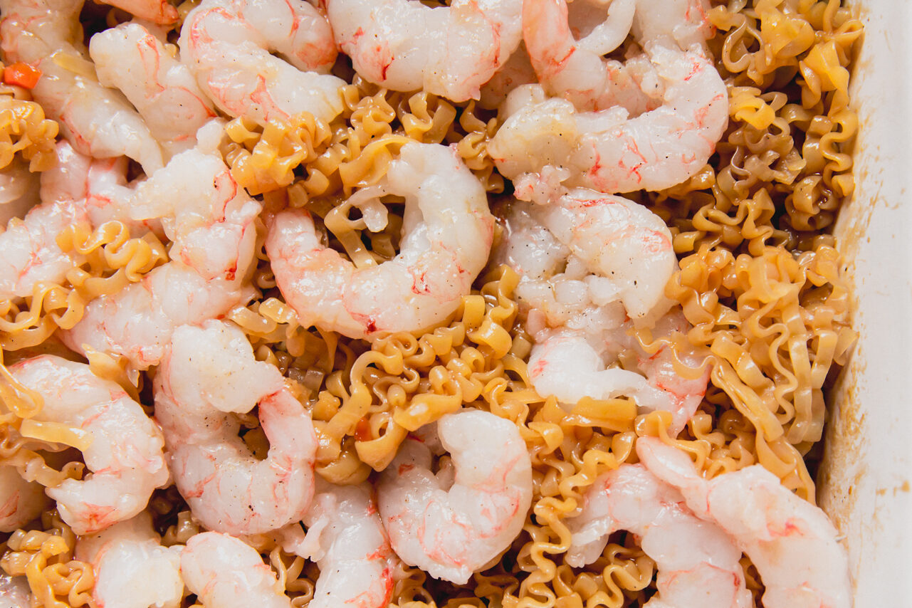 A white casserole dish with raw shrimp on top of a ramen and vegetable mixture.