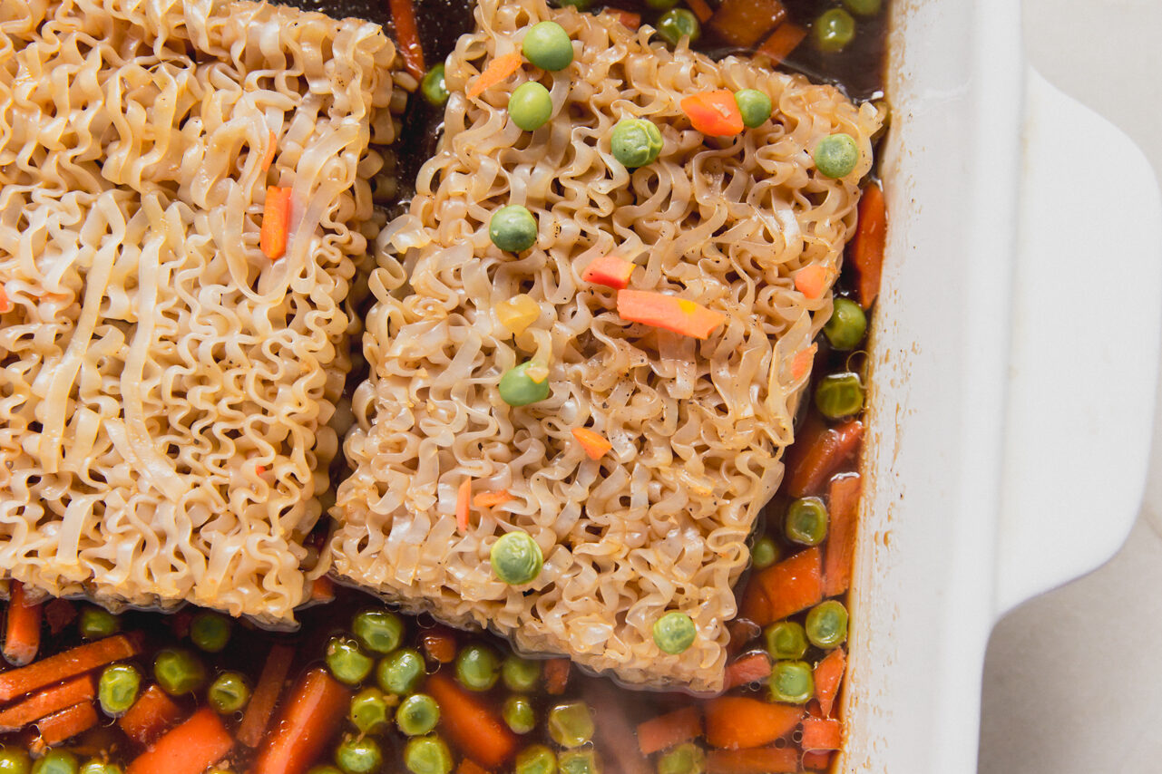 A white casserole dish with mixed vegetable and sauce topped with two ramen bricks.