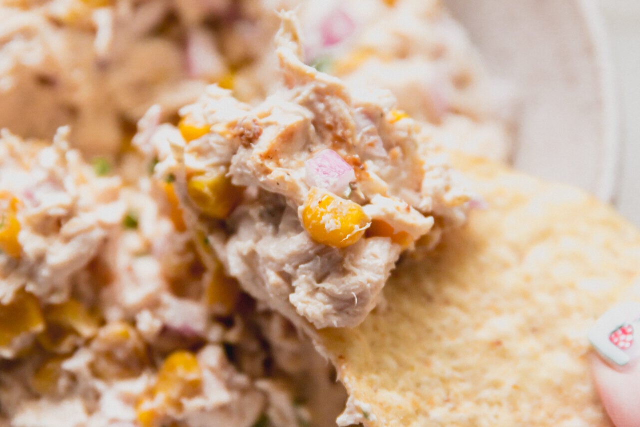 A chip taking a chunk of street corn chicken salad from the bowl.