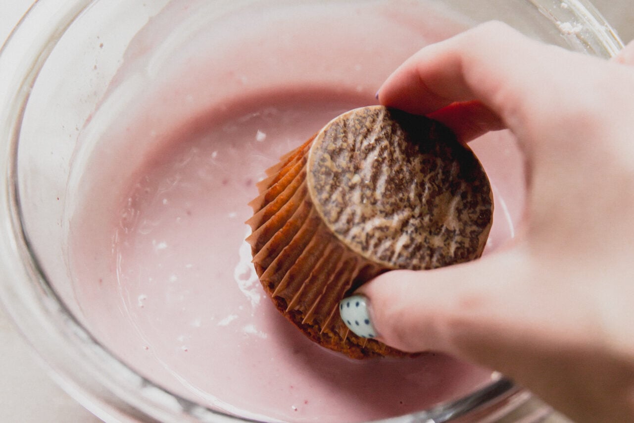 A hand dipping a high protein muffin into the greek yogurt icing in a glass bowl.